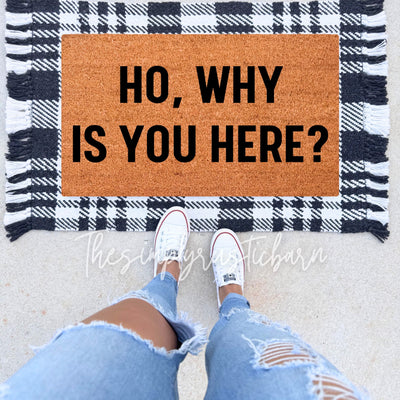 Funny doormat with bold black text that reads “Ho Why Is You Here?” on a coir doormat, placed at the entrance of a home.