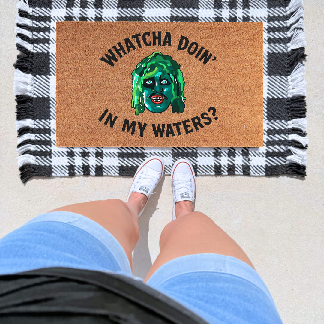 Funny horror inspired doormat with “Whatcha Doin’ In My Waters?” quote and old Gregg  design on a natural coir welcome mat layered over a black and white plaid rug.